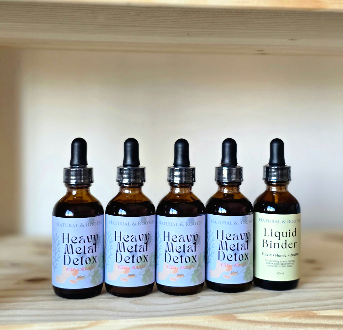Five amber glass dropper bottles of Natural & Root detox supplements arranged on a light wood shelf against a neutral beige background. Four bottles feature lavender labels reading 'Heavy Metal Detox' and the rightmost bottle has a pale yellow label reading 'Liquid Binder'.