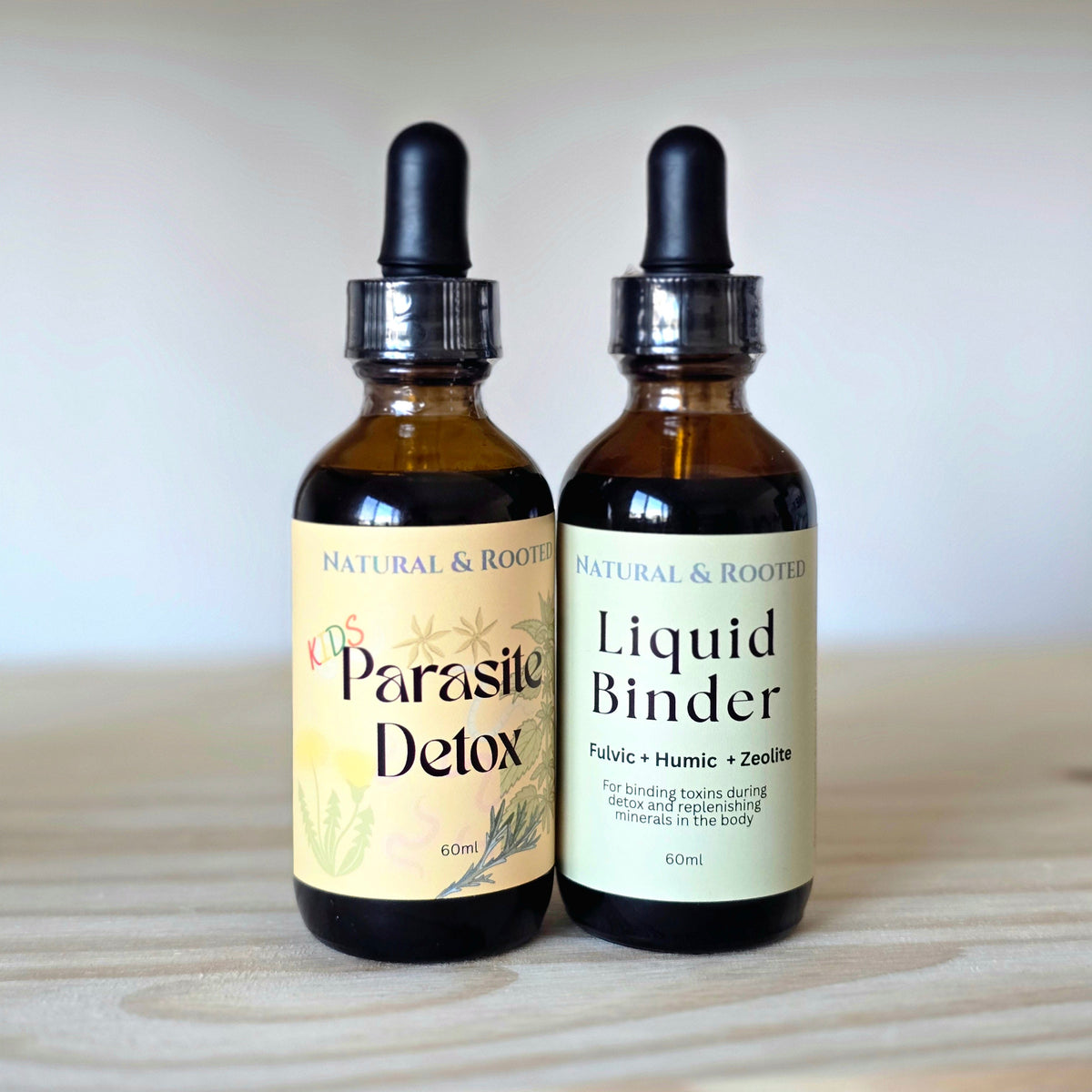 Two amber glass dropper bottles with black caps on a light wood surface against a soft beige background. The left bottle label reads 'Natural & Rooted' and 'Co-Enzyme Parasite Detox'.