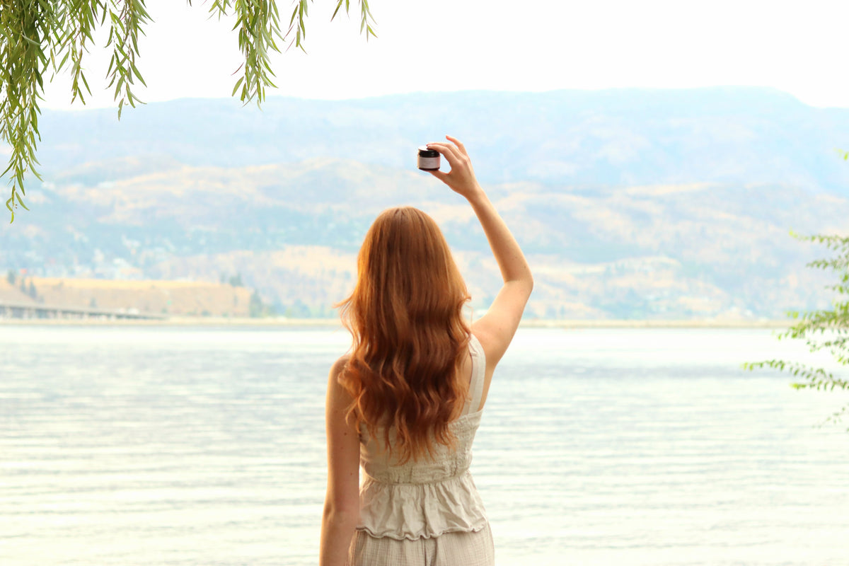 A woman with long red hair stands with her back to the camera, holding a small vial of product up against a scenic lake and mountain landscape.